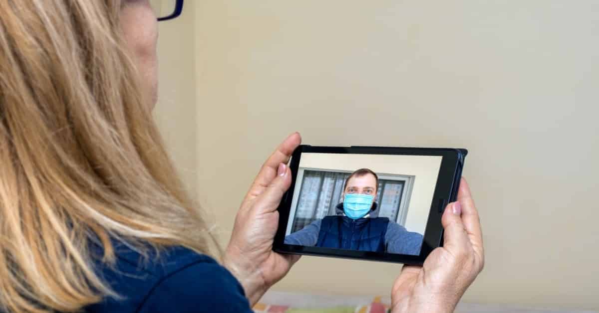 two people video calling, the one on the screen is masked, the other is holding the tablet.