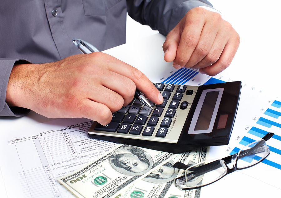 Image of a hand holding a pen typing on a calculator that is on top of three one hundred dollar bills fanned out over a spreadsheet.